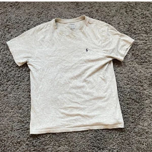 Beige t-shirt från Polo Ralph Lauren - Snygg grå t-shirt från Polo Ralph Lauren i nyskick med klassisk broderad logga på bröstet. Perfekt för en avslappnad stil. Kortärmad och tillverkad i mjukt material.