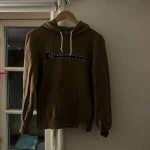 Brun hoodie från Champion - Säljer en brun hoodie från Champion med klassisk logga på bröstet. Tröjan har en bekväm passform med långa ärmar och en praktisk känguruficka framtill. Perfekt för en avslappnad stil.