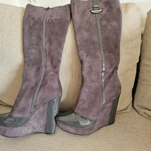 Grå mockastövlar med kilklack - Snygga lila/grå stövlar i mocka med kilklack och dragkedja. De har en glansig detalj vid foten och en dekorativ spänne upptill. Perfekta för att ge din outfit en elegant touch.