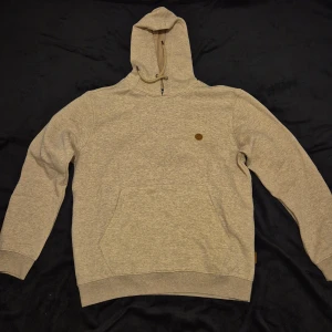 Hoodie med dragkedja - Säljer en stilren grå hoodie med dragkedja och en liten logotyp på bröstet. Perfekt för kyliga dagar med sin mjuka och bekväma passform. Den har långa ärmar och en praktisk ficka framtill.