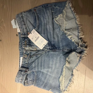 Jeansshorts - Snygga blå jeansshorts från Zara med fransiga kanter och en cool, sliten look. Perfekta för en avslappnad stil. De har en klassisk femficksdesign och knappgylf.