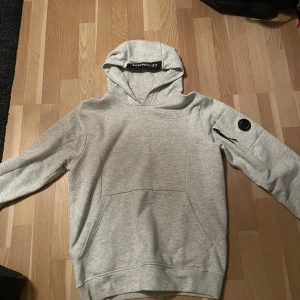 Grå hoodie från C.P. Company - Säljer en stilren grå hoodie från C.P. Company. Tröjan har en praktisk ficka på ärmen med en rund detalj och en klassisk huva. Perfekt för en avslappnad look.