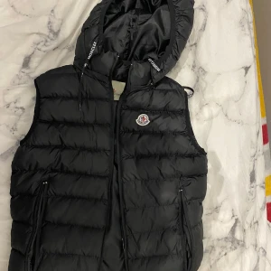 Svart dunväst från Moncler - Snygg svart dunväst från Moncler med huva och dragkedja. Västen har Moncler-logga på bröstet och tryck på insidan. Perfekt för kyliga dagar.