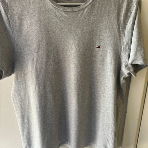Grå t-shirt från Tommy Hilfiger - Säljer en stilren grå t-shirt från Tommy Hilfiger. Den har en liten broderad logga på bröstet och är perfekt för en avslappnad look. T-shirten är i bomull och har en normal passform. Perfekt för vardagsbruk!