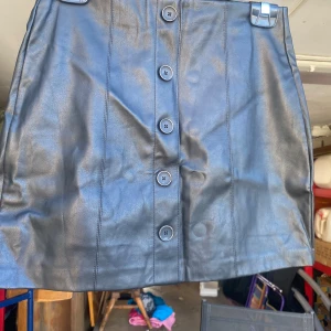 Svart kjol i skinnimitation från Pull&Bear - Säljer en snygg svart kjol i skinnimitation från Pull&Bear. Kjolen har en rad med knappar framtill som ger en cool look. Perfekt för både vardag och fest! 🖤