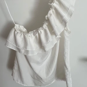 One shoulder blus - En superfin vit one-shoulder blus med fina dekorationer upptill. Köpte på Plick men har inte haft användning för den. Står ingen storlek men passar mig som har S/M💗Passar perfekt att ha i skolan och till fest.💗skriv för fler bilder eller frågor😃