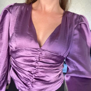 Lila satinblus från IVIVI - Säljer en superfin lila satinblus från IVIVI. Den har en v-ringad design med långa ärmar och snygg rynkdetalj framtill. Perfekt för både fest och vardag! 💜Ny med lappar kvar 