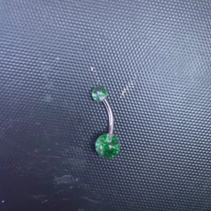 Grön navelpiercing med glittriga stenar - Snygg navelpiercing med en böjd stav i silverfärg och två gröna glittriga stenar. Perfekt för att lägga till lite bling till din stil. Passar både till vardags och fest!