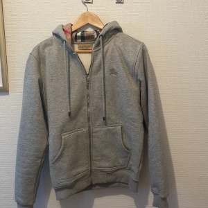 Grå hoodie från Burberry - Säljer en snygg och stilren grå hoodie från Burberry. Den har en dragkedja framtill och en klassisk huva med det ikoniska Burberry mönstret på insidan. Perfekt för både vardag och lite finare tillfällen. Den är perfekt skick och superbekväm att ha på