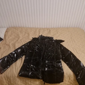 Guess Jacka - Säljer en snygg svart pufferjacka från Guess. Den är glansig och har en dragkedja framtill. Jackan är vindtät och perfekt för kyliga dagar. Den har långa ärmar och en huva för extra skydd mot väder och vind. Perfekt för både höst och vinter!