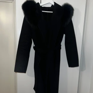 Svart lång kappa med äkta päls - Svart lång kappa med äkta päls. Köpt för mer än 1 år sen från butik Garoff men använts sparsamt. Detta är den långa modellen. I bra skick, inga hål osv. Storlek S/M. Går att ta av och på pälsen. Ull/ cashmere. 