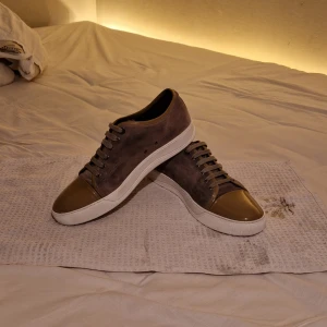 Lanvin cap toe grå/brun - Säljer ett par snygga sneakers från Lanvin i grå mocka med en glansig beige cap-toe. Skorna har en vit sula och snörning i matchande färg. Bara att höra av sig vid fundering eller prisförslag