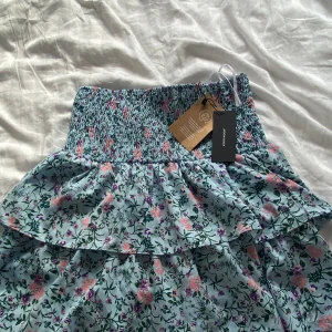 Blommig kjol från Vero Moda - Säljer en superfin blommig kjol från Vero Moda i storlek M. Den är i en härlig blå färg med rosa och gröna blommor. Som ni ser är den helt oanvänd och nypris är 300kr!