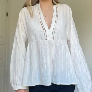 Vit blus från Zara - Säljer en superfin vit blus från Zara. Den har en v-ringad design med snygga spetsdetaljer längs halsen. Blusen är långärmad och har en luftig passform, perfekt för både vardag och fest. Tillverkad i ett lätt och bekvämt material.