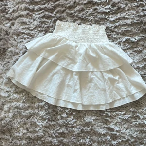 Vit volangkjol - Säljer en superfin vit kjol med volanger. Den har en elastisk midja som gör den bekväm och lätt att bära. Perfekt för sommardagar eller en festlig kväll. Kjolen är i ett lätt och luftigt material.