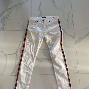 Vita byxor med ränder från Zara - Säljer ett par vita byxor från Zara med coola röda och svarta ränder längs sidorna tsm med fin grå Tshirt. Båda för 299kr. De är i storlek EUR 42 och har en klassisk femficksdesign med knapp och dragkedja. Perfekta för en avslappnad men stilren look!
