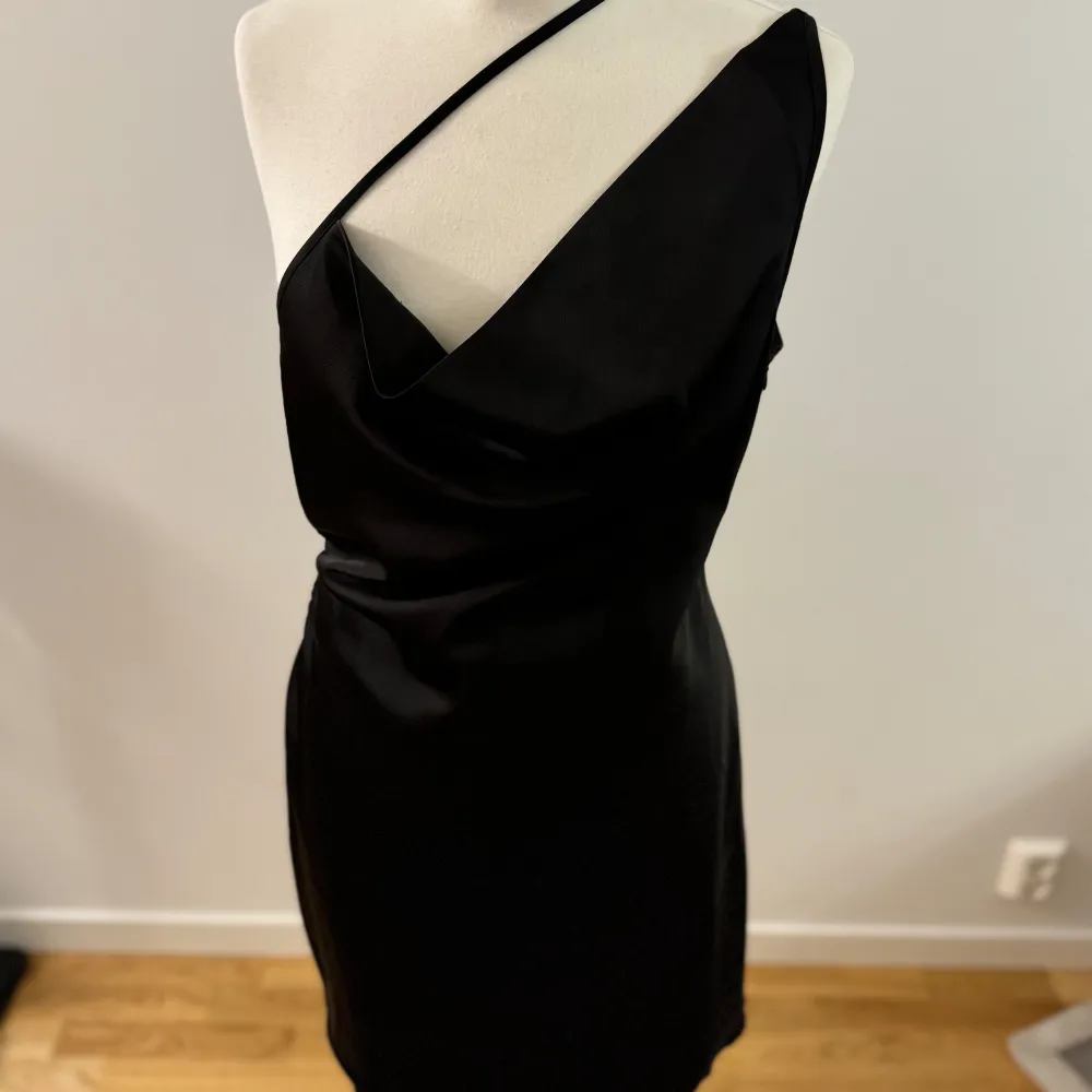 Säljer en elegant svart oneshoulder klänning från Chloé B. Den har en stilren design med en dragkedja i ryggen för enkel påklädning. Perfekt för festliga tillfällen!. Klänningar.