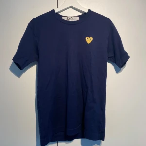 Commes Des Garçons T-shirt Navy - CDG T-shirt i Marinblå färg med gult hjärta. Storlek M men liten i storleken så passar S. Använd men i okej skick utan större defekter. Stilsäker & klassisk. Nypris 900kr på NK. Kan kombineras med andra plagg för bättre pris. Hörs i DM! 