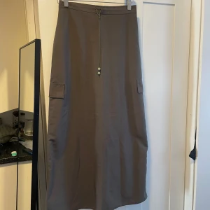 Brun långkjol med fickor - Säljer en snygg armegrön långkjol med praktiska fickor på sidorna. Kjolen har en enkel och stilren design med en dekorativ snörning framtill. Perfekt för höst och vår, och kan enkelt matchas med olika toppar för en avslappnad look.