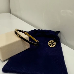 Tory Burch Svart & Guld Emaljerat Armband - Använd bara ett fåtal gånger!  Elegant svart och guld emaljerat armband med Tory Burch-logga. Ett mångsidigt tillbehör för alla garderober. • Svart och guld emalj • Tory Burch-logga • Hängslenstängning • Högkvalitativ finish • Lättviktsdesign