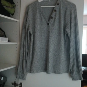 Grå tröja med knappar - Säljer en mysig grå tröja med v-ringning och dekorativa knappar längs halsen. Tröjan har långa ärmar och ett ribbat mönster vid ärmarna. Perfekt för kyliga dagar!