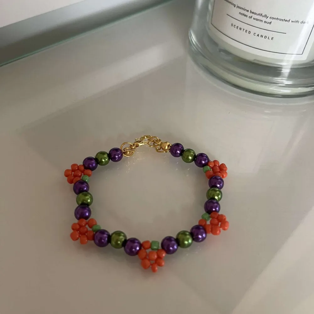 Säljer ett supercoolt armband med pärlor i lila, grönt och orange. Armbandet har små orange blommor som ger det en unik och lekfull stil. Perfekt för att liva upp vilken outfit som helst!. Accessoarer.