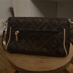 Mönstrad axelväska från Grazia Pirelli - Säljer en snygg och stilren axelväska från Grazia Pirelli. Väskan är mörkbrun med ett elegant mönster och har detaljer i guld. Den har en praktisk dragkedja framtill och en justerbar axelrem. Insidan är fodrad med ett ljust tyg med logotypmönster. Perfekt för både vardag och fest!