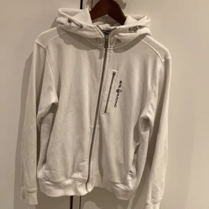 Vit hoodie från Sail Racing - Säljer en stilren vit hoodie från Sail Racing i storlek S. Den har en dragkedja framtill och en praktisk ficka med dragkedja på bröstet. Perfekt för både vardag och träning. Huvan har justerbara snören för bästa passform. Skön och bekväm att ha på sig!