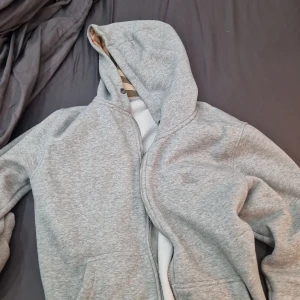 Grå hoodie från Burberry - Säljer en snygg grå hoodie från Burberry i mycket bra skick. Den har en klassisk rutig insida i huvan och dragkedja framtill. Perfekt för höst och vår, och den är supermjuk och bekväm. Passar perfekt för en casual look eller när du vill ha något stilrent och enkelt. 🧥✨