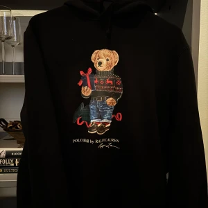 Svart hoodie med Polo Bear från Ralph Lauren - Säljer en svart hoodie från Ralph Lauren med den ikoniska Polo Bear på framsidan. Björnen har en jultröja och håller en present, vilket gör den perfekt för vintern och julen. Hoodien är i bra skick och har en mysig passform. Perfekt för att hålla sig varm och stilren under kalla dagar! Priset är ej hugget i sten, så vid smidig affär så går priset att sänkas👍🏻