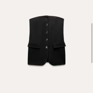 Svart korsettliknande topp - Snygg svart korsettliknande topp med knappar framtill. Den är axelbandslös och har en stilren design som passar perfekt för festliga tillfällen. Toppen har en figurnära passform och är tillverkad i ett elegant material. Orginalpris: 500kr