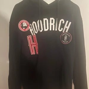 Svart hoodie från Hoodrich - Säljer en svart hoodie från Hoodrich med coola patches och tryck i vitt och rött. Den har en klassisk känguruficka och justerbar huva med dragsko. Perfekt för en avslappnad stil eller streetwear-look. Passar bra till både vardag och chill.
