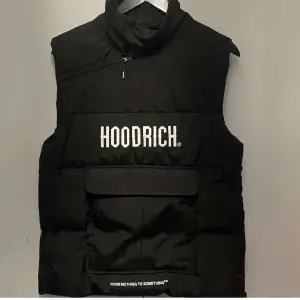 Säljer en svart dunväst från Hoodrich i mycket bra skick. Västen har en dragkedja och en stor ficka framtill med texten 'FROM NOTHING TO SOMETHING'. Perfekt för höst och vinter, och den är helt ny allltså oanvänd. Passar bra för både vardag och fest.  Nypris:1000kr och säljs inte längre! Pris går att diskutera vid snabbaffär.