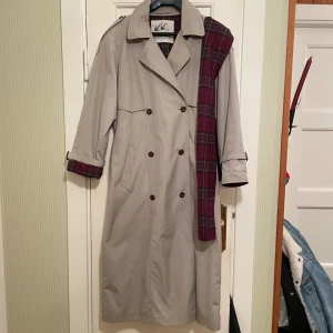 Beige trenchcoat från Scandinavian Design - Perfekt höstkappa! Stilren gråbeige trenchcoat från Scandinavian Design. Den har en klassisk dubbelknäppning och ett bälte i midjan för en snygg passform. Insidan är varmfodrad med ett röd- och blå-rutigt mönster som ger en cool kontrast. Perfekt för höst och vår, men funkar även på vintern. 120 cm lång. Matchande halsduk medföljer.