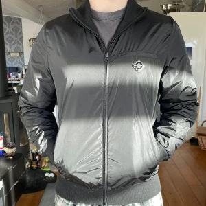 Svart vårjacka  - Svart vårjacka från J.Lindeberg, med två jack sidor (dvs jackans inner och yttertyg kan användas som jacka, ena sidan är i blankt polyester och andra sidan är i ull & polyester tyg). Den har en ribbad krage vid halsen och ribbade ärmmuddar. Perfekt som vår, sommar och höst jacka. 2600kr nypris