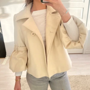 Beige kappa med puffärmar - Säljer en superfin beige kappa med unika puffärmar och stora knappar framtill. Perfekt för höst och vår, den ger en chic och stilren look. Jackan har en lös passform och är i mycket bra skick. Perfekt för att lyfta vilken outfit som helst!