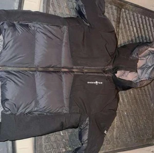 Svart dunjacka från Sail Racing - Säljer en svart dunjacka från Sail Racing i bra skick. Jackan har en lång dragkedja och är perfekt för kalla vinterdagar. Den är långärmad och har en stilren design med en huva för extra värme. Perfekt för både vardag och utomhusaktiviteter!