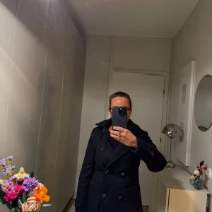 Marinblå kappa från Bien Bleu med luva - Säljer en stilren marinblå kappa från Bien Bleu med luva. Den har en klassisk design med dubbelknäppning och ett snyggt röd- och vitrutigt foder. Perfekt för höst och vinter! 🖤