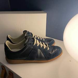 Säljer ett par snygga blå sneakers från Maison Margiela i mycket bra skick. Skorna har en stilren design med beige snörning och en gummisula i ljusbrun färg. Perfekta för både vardag och festliga tillfällen. Kommer i originalkartong.  Box ingår. De är inprincip ny skick 9,5-10. Priset är inte hugget i sten. Ställ gärna frågor om ni har någon ?