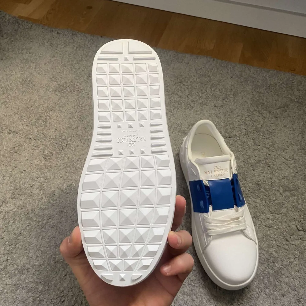 Säljer ett par snygga Valentino Garavani sneakers i vitt med blå detaljer. Skorna är i nytt skick och kommer med originalförpackning och dustbag. Perfekta för både vardag och fest! Storlek och kvitto finns med på bilden. Passa på att köpa dessa stilrena skor!. Skor.