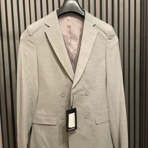 Grå kavaj från boohooMAN - Säljer en snygg grå kavaj från boohooMAN i nytt skick. Den har en dubbelknäppt design med fyra knappar och två fickor fram. Perfekt för både fest och formella tillfällen. Passar bra till vår och sommar. Kavajen är i storlek 34 och har en stilren look.