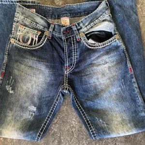 True Religion jeans  - Säljer ett par snygga True Religions jeans i storlek 33. De är i en klassisk blå färg med coola slitningar. Jeansen har en normal passform och är tillverkade i 100% bomull, vilket gör de båda bekväma och hållbara. De är köpte från Vintage & Friends för 1.5.