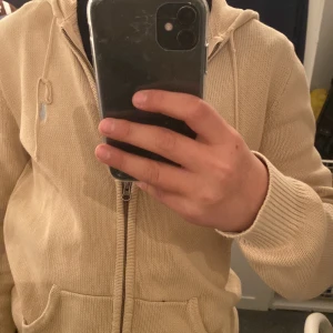 Beige stickad hoodie med dragkedja - Säljer en snygg beige stickad hoodie från Ralph Lauren. Den har en dragkedja framtill och en broderad logga på bröstet. Perfekt för höst och vår, med långa ärmar och en mysig huva. Passar både till vardags och lite mer avslappnade tillfällen.