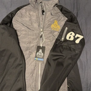 Grå och svart jacka från Chrixton - Säljer en snygg grå och svart jacka från Chrixton i storlek M. Jackan har en dragkedja framtill och en cool detalj med siffran 67 på ärmen. Perfekt för höst och vår med sitt stilrena utseende och bekväma passform. 