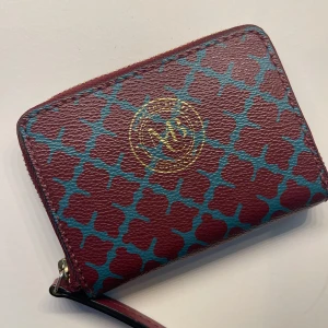 Malene Birger Mönstrad plånbok med dragkedja - Säljer en snygg plånbok i mycket bra skick med ett unikt mönster i rött och blått. Plånboken har en praktisk dragkedja och är perfekt för att hålla ordning på kort och kontanter. Den är kompakt och lätt att ta med sig överallt. Frakt betalar av köparen.