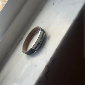 Svart och silver armband från Boss - Snyggt armband från Boss i svart och silver. Det har en stilren design med en metallplatta där det står 'BOSS'. Perfekt för att ge en elegant touch till din outfit. Armbandet är i mycket bra skick och passar både till vardags och fest.