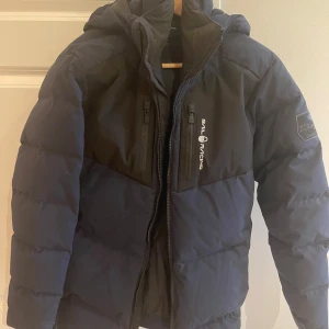 Svart dunjacka från Sail Racing - Säljer en svart dunjacka från Sail Racing i mycket bra skick. Jackan är perfekt för kalla vinterdagar med sitt 500 fill power dun. Den har en dragkedja framtill och två bröstfickor med dragkedjor. Jackan är långärmad och har en stilren design med en liten logga på bröstet. Perfekt för både vardag och utomhusaktiviteter!