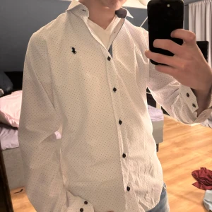 Vit skjorta från Polo Ralph Lauren - Säljer en snygg vit skjorta från Polo Ralph Lauren i nytt skick. Skjortan har ett diskret prickigt mönster och är långärmad med svarta knappar. Perfekt för både vardag och fest! 🕺