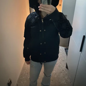 Svart pufferjacka från Glorious Gangsta - Säljer en snygg svart pufferjacka från Glorious Gangsta. Jackan har ett coolt geometriskt mönster på ärmar och huva, samt dragkedja och knappar framtill. Perfekt för höst och vinter med sin varma och stiliga design.