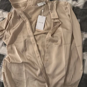 Från VILA - Säljer en elegant beige blus från Vila, perfekt för både vardag och fest. Blusen är tillverkad av återvunnen polyester och har en klassisk krage med knappar framtill. Den har långa ärmar med knappdetaljer vid ärmsluten. Superfin och miljövänlig! 🌿